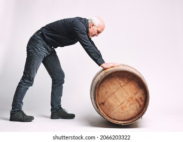 Senior Man Rolling An Old Oak Wine Or Beer Barrel In Front Of Him Along The Floor In A Full Length Side View On White