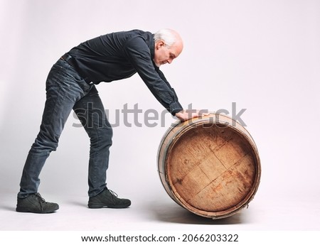 Senior man rolling an old oak wine or beer barrel in front of him along the floor in a full length side view on white