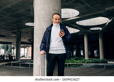 Senior Man With Rolling Suitcase Standing Outdoors Looking At Mobile Phone