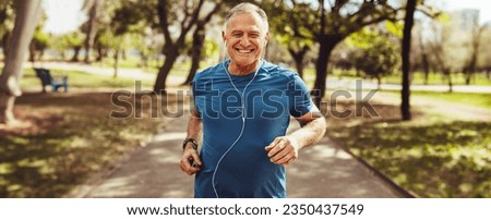 Senior man running in a park with a smile on his face and earphones on. Elderly man embracing physical fitness, caring for his health and wellbeing and longevity in his golden years.