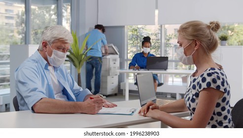 Senior Man In Safety Mask Having A Business Meeting And Signing Contract. Mature Client Sign Lease Or Purchase Agreement Given By Bank Manager In Protective Mask