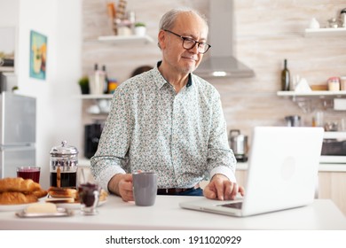 Senior Man Searching Online Recipe Using Laptop In Modern Kitchen During Breakfast. Elderly Retired Person Working From Home, Telecommuting Using Remote Internet Job Online Communication