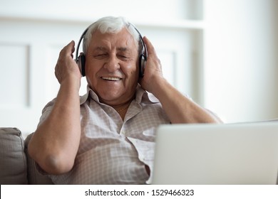 Senior man seated on sofa in living room hold laptop on lap wearing headphones listens favourite track having nostalgic mood enjoys songs of his youth, older generation using modern technology concept