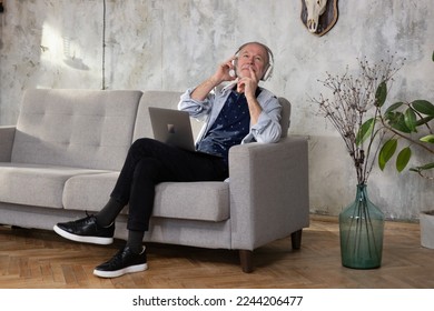 Senior man seated on sofa in living room hold laptop on lap wearing headphones listens favourite track having nostalgic mood enjoys songs of his youth, older generation using modern technology concept
