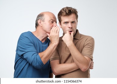 Senior man sharing secret or whispering gossips into his son ear, who is shocked and looking at camera with shocked expression on his face. Telling family secret concept