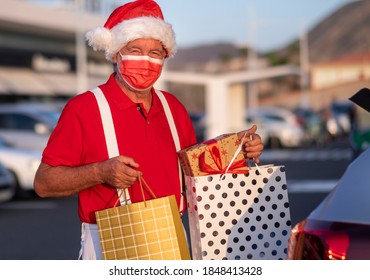 A Senior Man Shopping For Christmas In A Santa Hat And Suspenders, Wearing A Medical Mask To Avoid Covid-19 Coronavirus Contagion, Puts Shopping Bags With Gifts In The Car Trunk In The Mall Parking