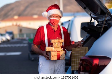 A Senior Man Shopping For Christmas In A Santa Hat And Suspenders, Wearing A Medical Mask To Avoid Covid Coronavirus Infection, Holding A Shopping Bag Full Of Gifts While Standing In The Mall Parking