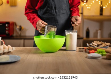 Senior man sifts flour into a bright green bowl while preparing dough for Christmas cookies. Baking utensils, eggs, and cookie cutters are arranged on the table, evoking a festive kitchen atmosphere. - Powered by Shutterstock