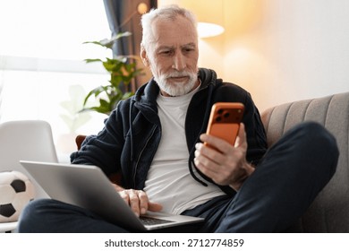 Senior man simultaneously using a smartphone and laptop, multitasking on a couch at home