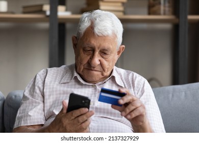 Senior Man Sit On Sofa At Home Holds Smartphone And Credit Card, Paying Remotely, Makes Instant Money Transfer. Older Gen Using Modern Tech, E-payments And E-bank Apps, Comfort Services Usage Concept