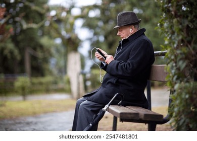 A senior man sits quietly on a park bench, engaged with his smartphone. He listens to music with earbuds, enjoying a peaceful moment outdoors in a green environment. - Powered by Shutterstock