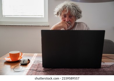 Senior Man Sitting At Domestic Dining Table Drinking Coffee And Using Laptop Computer For A Zoom Meeting With A Family.