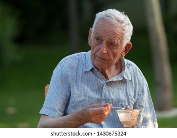 Senior Man Sitting In Garden And Eating Ice Cream From Glass Bowl, With Green Background