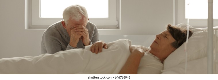 Senior Man Sitting Next To His Dying Wife's Hospital Bed And Crying