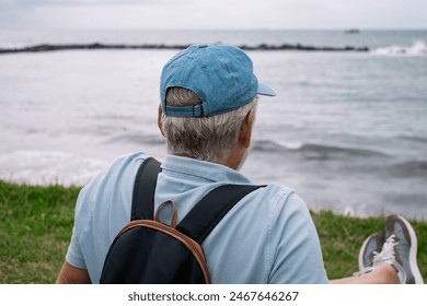 Senior man sitting on the grass at the beach in a cloudy day enjoying free time vacation or retirement. Bearded relaxed man wearing blue cap looking at the horizon over sea - Powered by Shutterstock