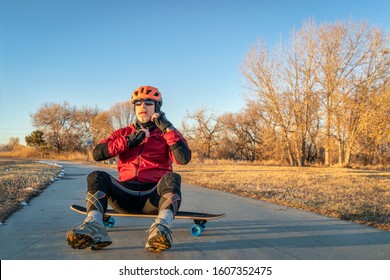 Senior Man Is Sitting On A Skateboard And Putting Helmet, A Paved Bike Trail In Winter Scenery In Northern Colorado - Boyd Lake State Park