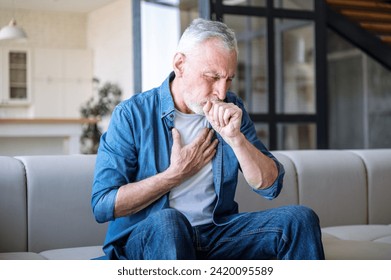 Senior man sitting on sofa at home and holding hand on chest. Male having asthma attack, difficulties with breathe, feeling severe pain or dyspnea
