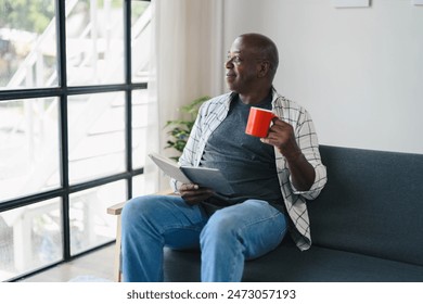 Senior man sitting on a sofa, holding a red coffee mug and a tablet, looking out the window in a modern living room. - Powered by Shutterstock