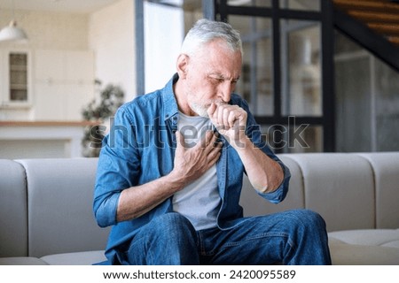 Senior man sitting on sofa at home and holding hand on chest. Male having asthma attack, difficulties with breathe, feeling severe pain or dyspnea