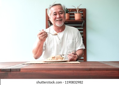 Senior Man Sitting Outside Eating Lunch On A Beautiful Day. Old Asian Male With White Beard Sitting On Wooden Bench With Wooden Table. Senior Home Service Concept.