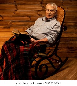 Senior Man Sitting In Rocking Chair In Homely Wooden Interior With Old Book