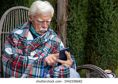 Senior Man Sitting In The Rocking Chair Reading Text Message On Smartphone. Mature Man Using His Smartphone On The Veranda. Portrait Of A Retired Man Reading A Message On His Cellular.