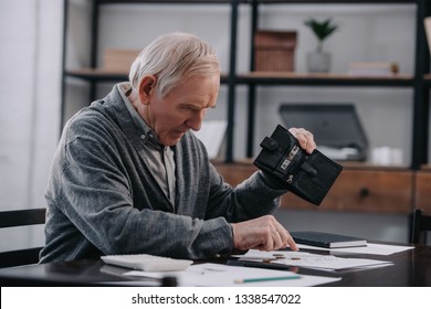 senior man sitting at table with paperwork, holding wallet and counting money at home