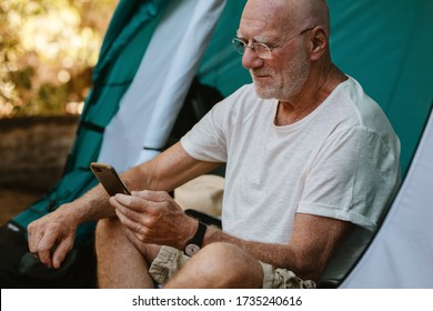 Senior man sitting in tent and using his mobile phone. Mature man camping in nature reading text message on his smartphone. - Powered by Shutterstock