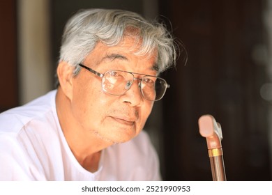 A senior man sitting thoughtfully while holding a walking cane, dressed in a white shirt and glasses. He appears to be in a peaceful, reflective moment, with natural sunlight highlighting his face.  - Powered by Shutterstock