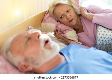 Senior Man Sleeping On Back In Bed And Snoring While Woman Is Awake And Annoyed Holding Pillow On Ears