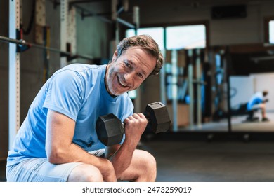 A senior man smiles as he sits on a bench and lifts a dumbbell. He is working his biceps in a gym. - Powered by Shutterstock