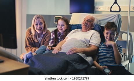 Senior man smiling and talking with daughter and grandchildren while lying on bed and watching TV in clinic ward
