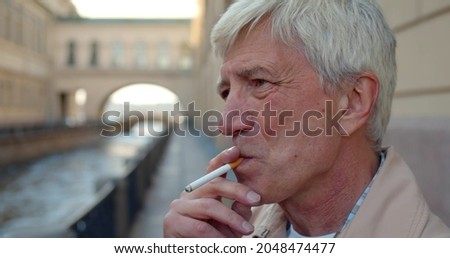 Senior man smoking cigarette outdoors in city. Close up portrait of aged male smoke outside near city river. Healthcare and bad habit concept