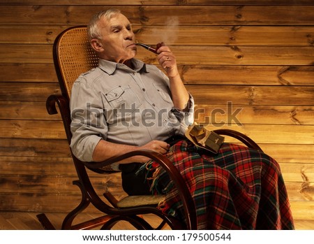 Senior man with smoking pipe sitting on rocking chair in homely wooden interior