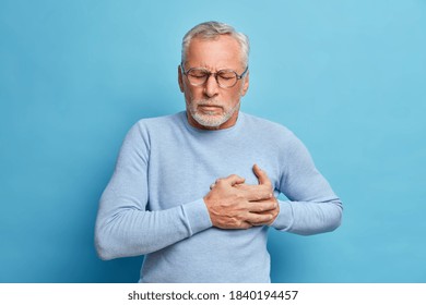 Senior man in spectacles presses hand to chest has heart attack suffers from unbearable pain closes eyes wears optical glasses poses against blue background. People age and problems with health