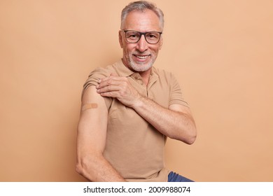 Senior man in spectacles shows plastered arm after getting coronavirus vaccine happy to feel safe and protected isolated over brown background. Health care during pandemic. Old people vaccination - Powered by Shutterstock