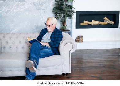 Senior Man Spends Leisure Time Reading Book Sitting On The Comfortable Couch At Cozy Home. A Gray-haired Older Man In Eyeglasses Immersed In Reading