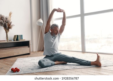 Senior man in sport clothing stretching while sitting on the floor near the window at home - Powered by Shutterstock
