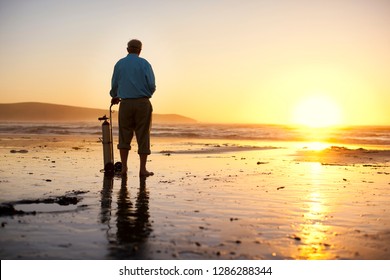 Senior Man Standing On A Beach With His Oxygen Tank At Sunset.