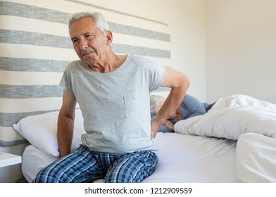 Senior Man Suffering From Back Pain At Home While Wife Sleeping On Bed. Old Man With Backache Having Difficulty In Getting Up From Bed. Suffering From Backache And Sitting On Bed In The Morning.