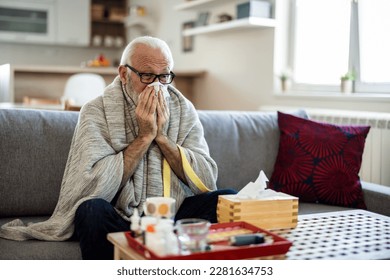 Senior man suffering from flu while sitting wrapped in a blanket on the sofa at home. Elderly Man blowing his nose while lying sick in bed at home. Man with a cold lying in sofa holding tissues - Powered by Shutterstock