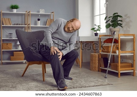 Senior man suffers from strong pain in his injured leg. Retired old man sitting on his chair at home and holding his aching knee with an expression of pain on his face. Old age health problems concept