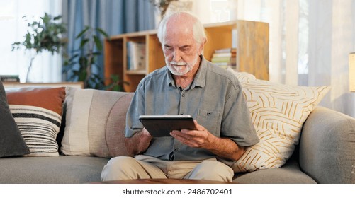 Senior man, tablet and typing on sofa for web surfing with retirement, memory game or connection in home. Elderly person, tech and relax with reading ebook, online puzzle or internet scroll in lounge - Powered by Shutterstock