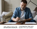 Senior man taking notes in notebook while sitting on the couch, clever gray-haired elderly man writing down thoughts and ideas in notebook
