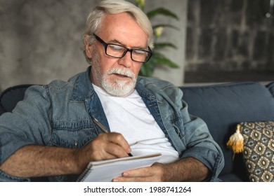 Senior man taking notes in notebook while sitting on the couch, clever gray-haired elderly man writing down thoughts and ideas in notebook - Powered by Shutterstock