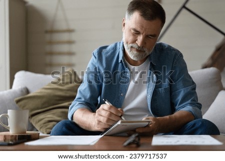 Senior man taking notes in notebook while sitting on the couch, clever gray-haired elderly man writing down thoughts and ideas in notebook