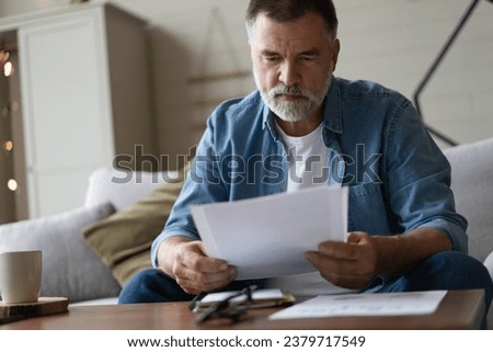Senior man taking notes in notebook while sitting on the couch, clever gray-haired elderly man writing down thoughts and ideas in notebook