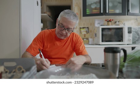 Senior man taking notes while seated in kitchen. A 70s male caucasian person writes information of paper with pen