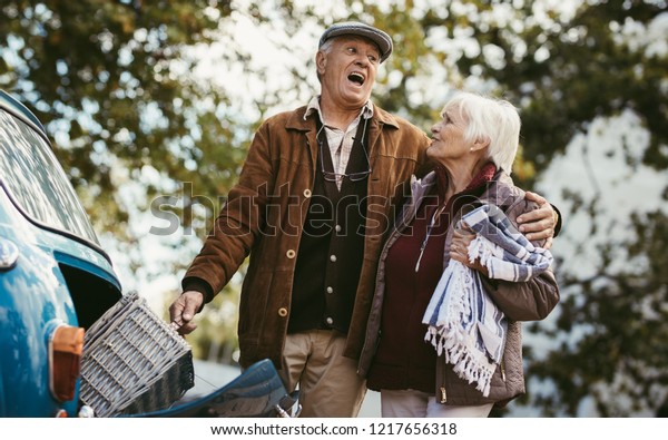 Senior man taking out\
the picnic basket out of car trunk and making expression as it is\
very heavy, holding his wife. Senior couple going on picnic on a\
winter day.