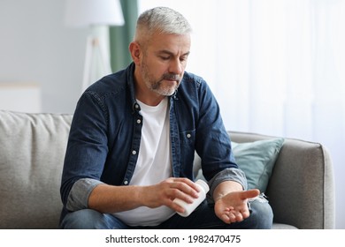 Senior Man Taking Pills While Sitting On Couch At Home, Holding White Jar With Treatment, Copy Space. Grey-haired Elderly Man Using Supplements Or Vitamins. Healthy Lifestyle In Senior Age Concept